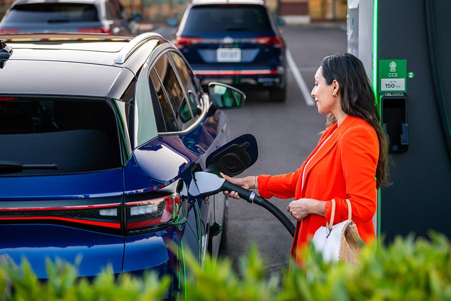 A well-dressed woman, parked at charging station stands on the passenger side of an ID.4 in Dusk Blue Metallic as she plugs in the car.